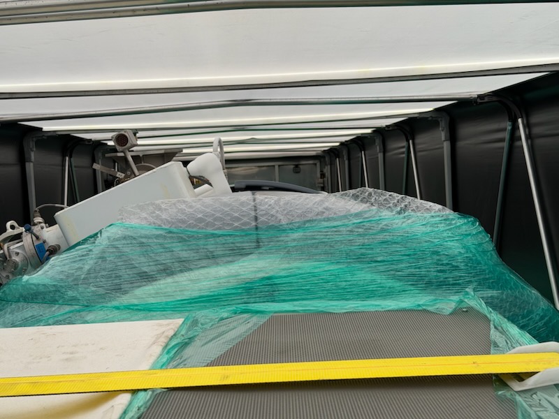 Interior view of a Conestoga trailer showing the roof bows, curtain rails, and a yellow strap running across shrink-wrapped machinery on the deck