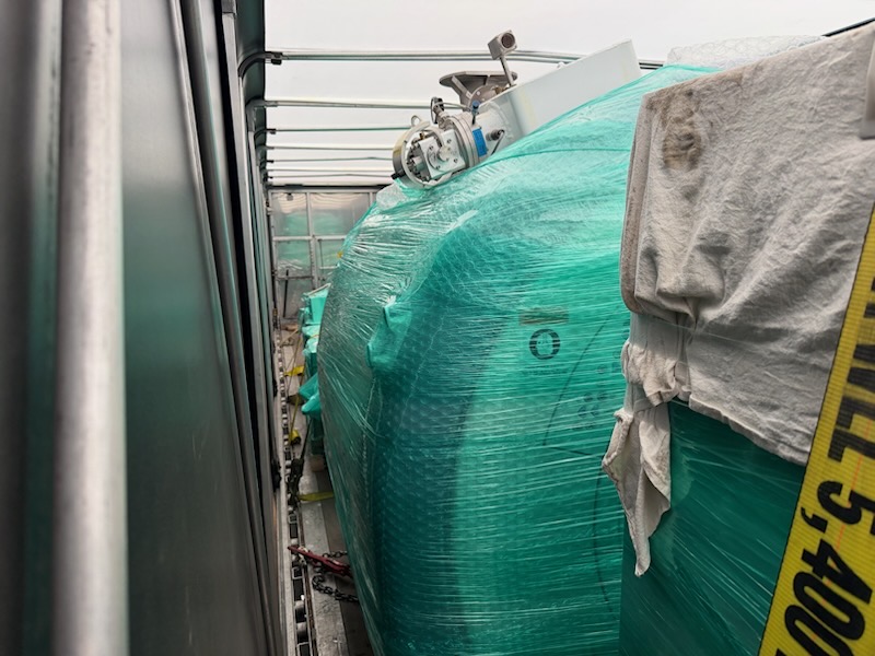 Shrink-wrapped industrial machinery inside a Conestoga trailer secured with a yellow strap marked working load limit 5,400 lbs and chain tie-downs at the deck