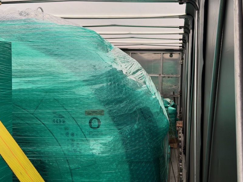 Green shrink-wrapped equipment loaded on the deck of a Conestoga curtain-side trailer with the side curtain open