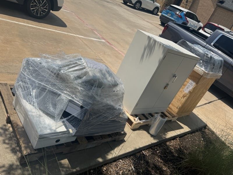 Palletized, shrink-wrapped industrial equipment and a metal control cabinet set down for curbside unloading