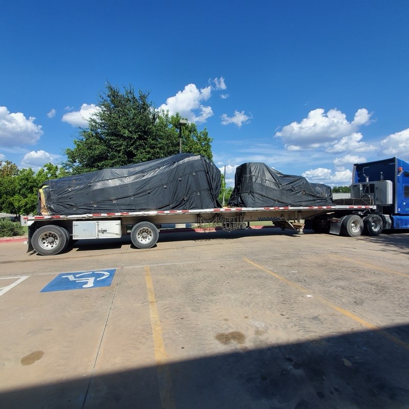 Two tarped machinery loads secured on a flatbed trailer behind a blue Kenworth tractor, tarps tensioned with no loose edges
