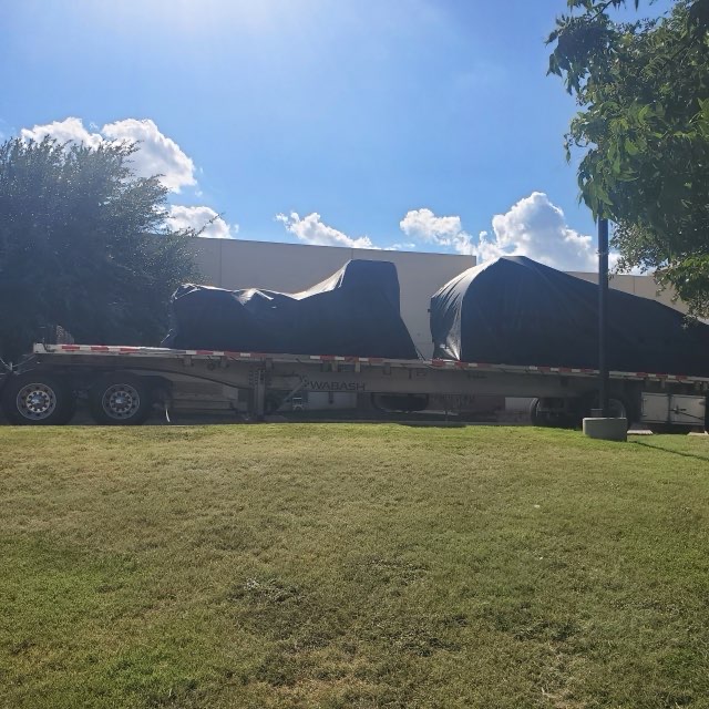 Flatbed trailer loaded with two black-tarped machinery loads staged outside a warehouse before departure